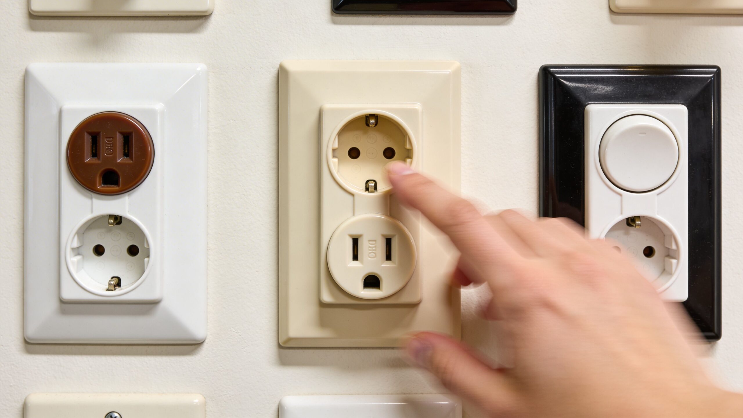 En hånd rører ved et gammelt dæksel til et lampeudtag og en stikkontakt på en hvid væg.