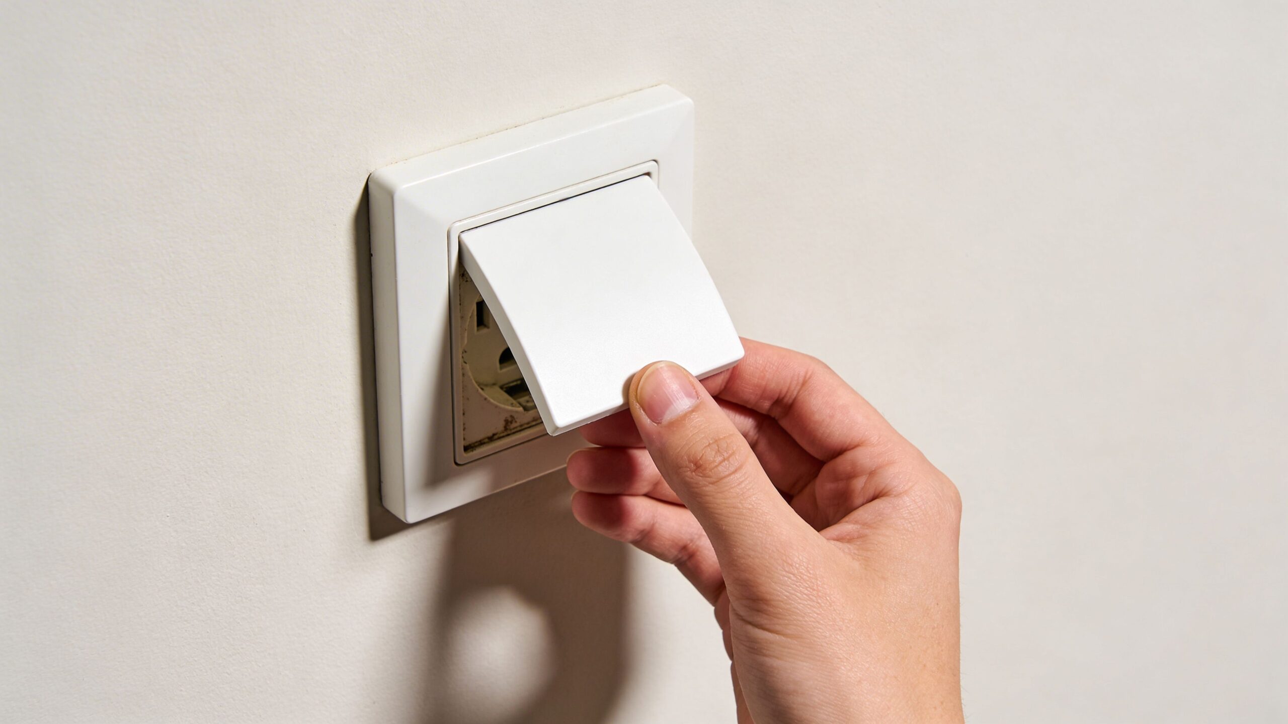 En hånd der løfter dækslet af et gammelt lampeudtag monteret på en hvid væg i hjemmet